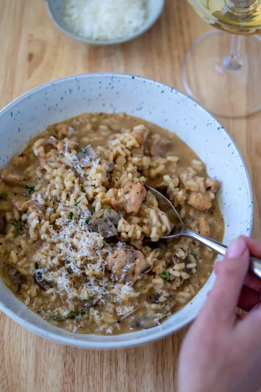 Hand holding a spoonful of creamy chicken and mushroom risotto in a speckled bowl, topped with grated Parmesan, with a bowl of cheese and a glass of white wine in the background.