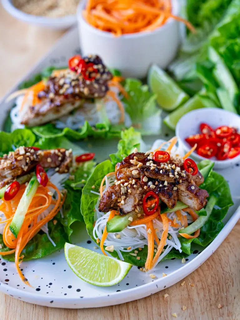 Sideways view of a full platter featuring hoisin chicken lettuce cups with vermicelli noodles, julienned vegetables, sliced red chili, crushed peanuts, and lime wedges, surrounded by fresh garnishes.