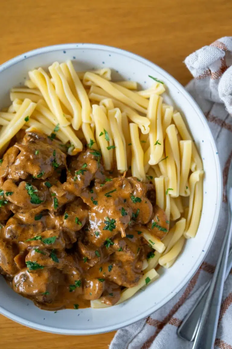 Top view of a white speckled bowl filled with slow-cooked beef stroganoff and pasta, showcasing tender beef in a rich creamy sauce alongside twisted pasta noodles, garnished with chopped parsley and served on a wooden table with a checkered napkin and silver cutlery.