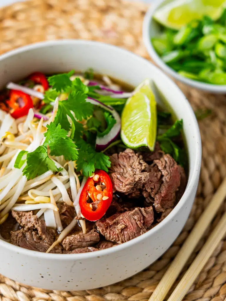 Overhead angled shot of Cheat's Beef Pho, showcasing slices of beef, rice noodles, fresh herbs, and lime in a rich broth.