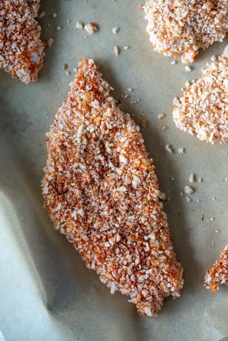 Uncooked crumbed chicken tenders on a parchment-lined baking tray, ready for the oven.