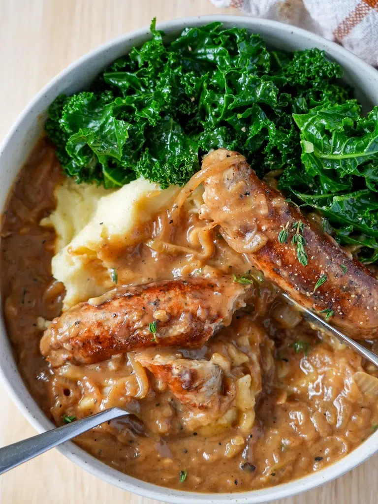 A bowl of Sausages served with homemade onion gravy, garnished with fresh herbs, and accompanied by mashed potatoes