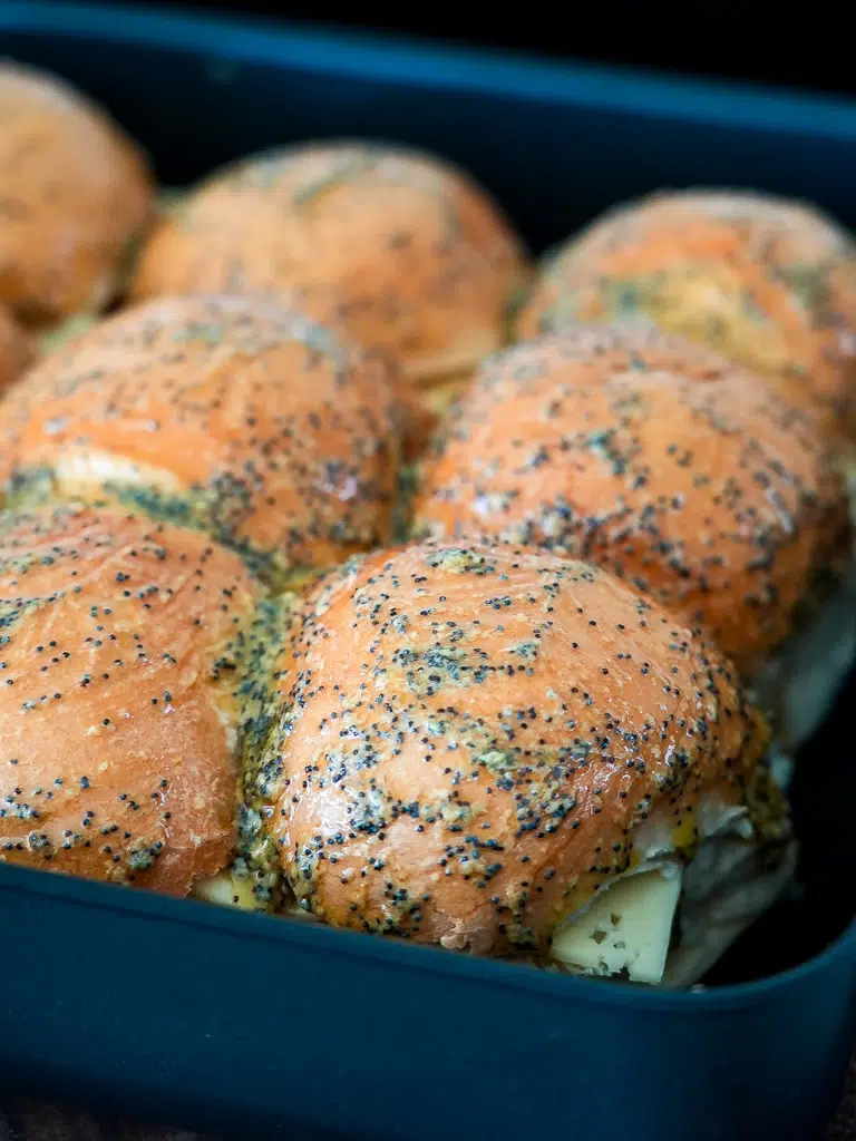 Close-up of baked ham and cheese sliders in a pan, topped with a shiny, crispy glaze of buttery honey garlic mustard and sprinkled with poppy seeds.