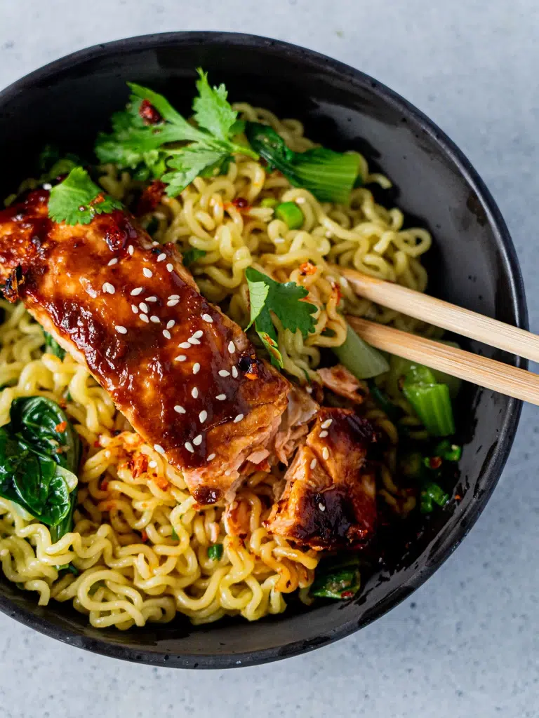 A bowl of miso-glazed salmon ramen with chopsticks pulling up noodles.