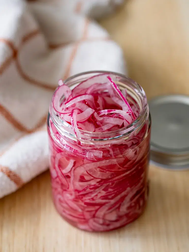 Open jar of freshly made pickled red onions on a wooden surface with a kitchen towel in the background.