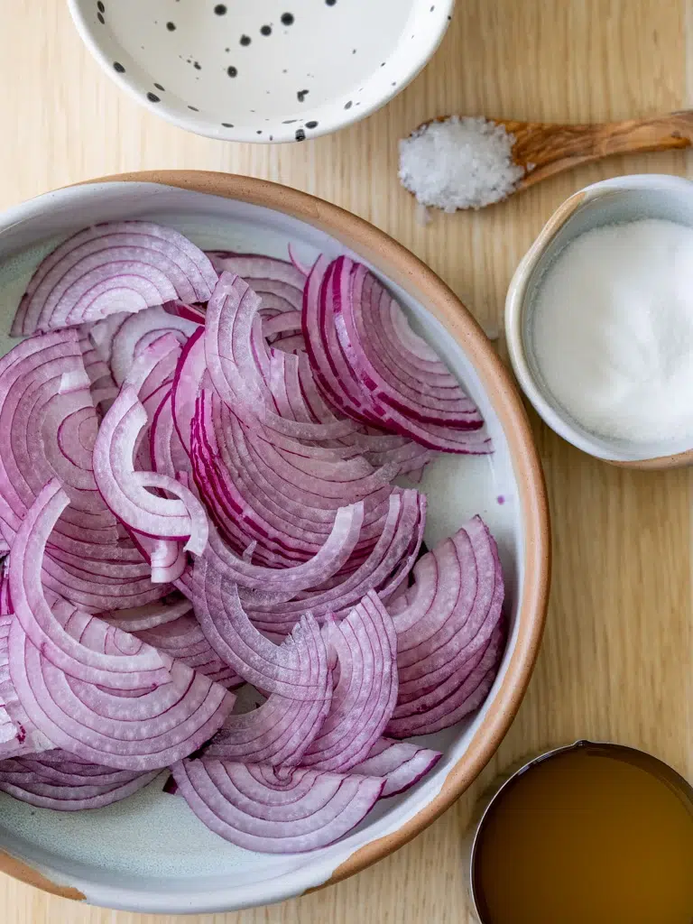 Sliced red onions in a bowl with salt, sugar, vinegar, and a speckled bowl prepared for pickling.