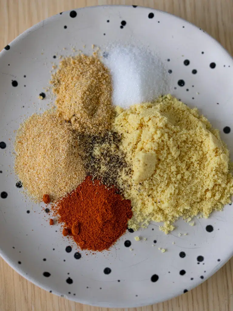 A plate with individual piles of spices for homemade chicken salt seasoning, including paprika, garlic powder, onion powder, salt, pepper, and chicken bouillon powder.