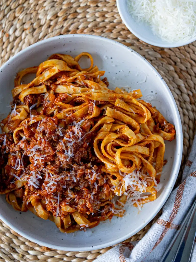 Top-down view of hidden vegetable Bolognese fettuccine served in a white bowl, topped with grated Parmesan, with extra cheese in a small dish on the side