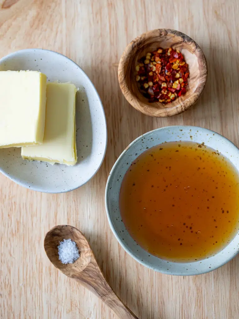 Flat lay of ingredients for hot honey sauce, including butter slices, crushed red pepper flakes, honey in a bowl, and a wooden spoon with salt.