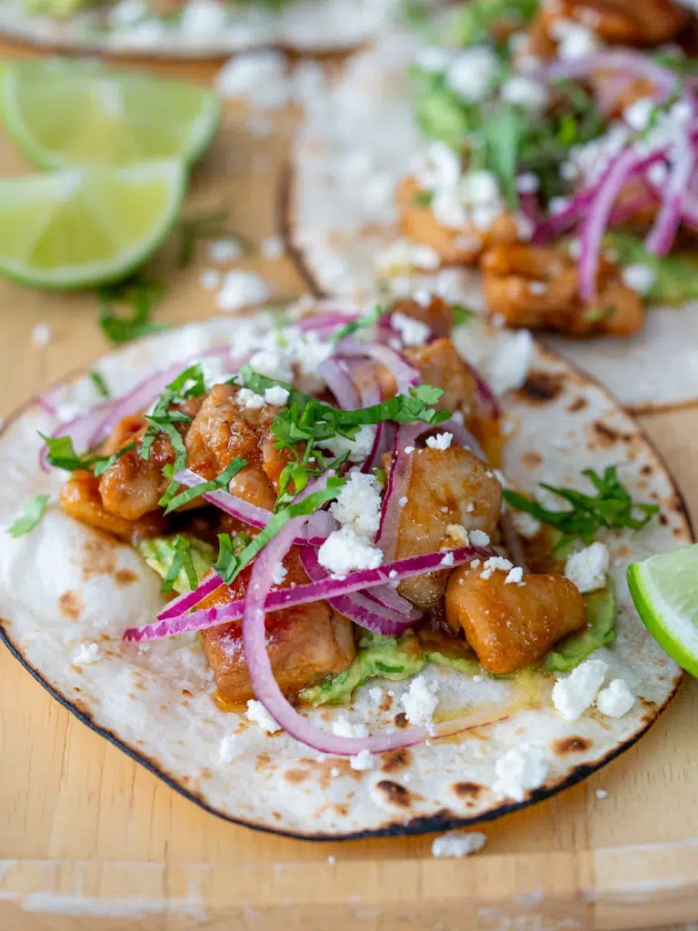 "Close-up of sticky lime honey glazed chicken taco with guacamole, pickled red onions, crumbled cheese, and chopped herbs on a toasted flour tortilla, served with lime wedge