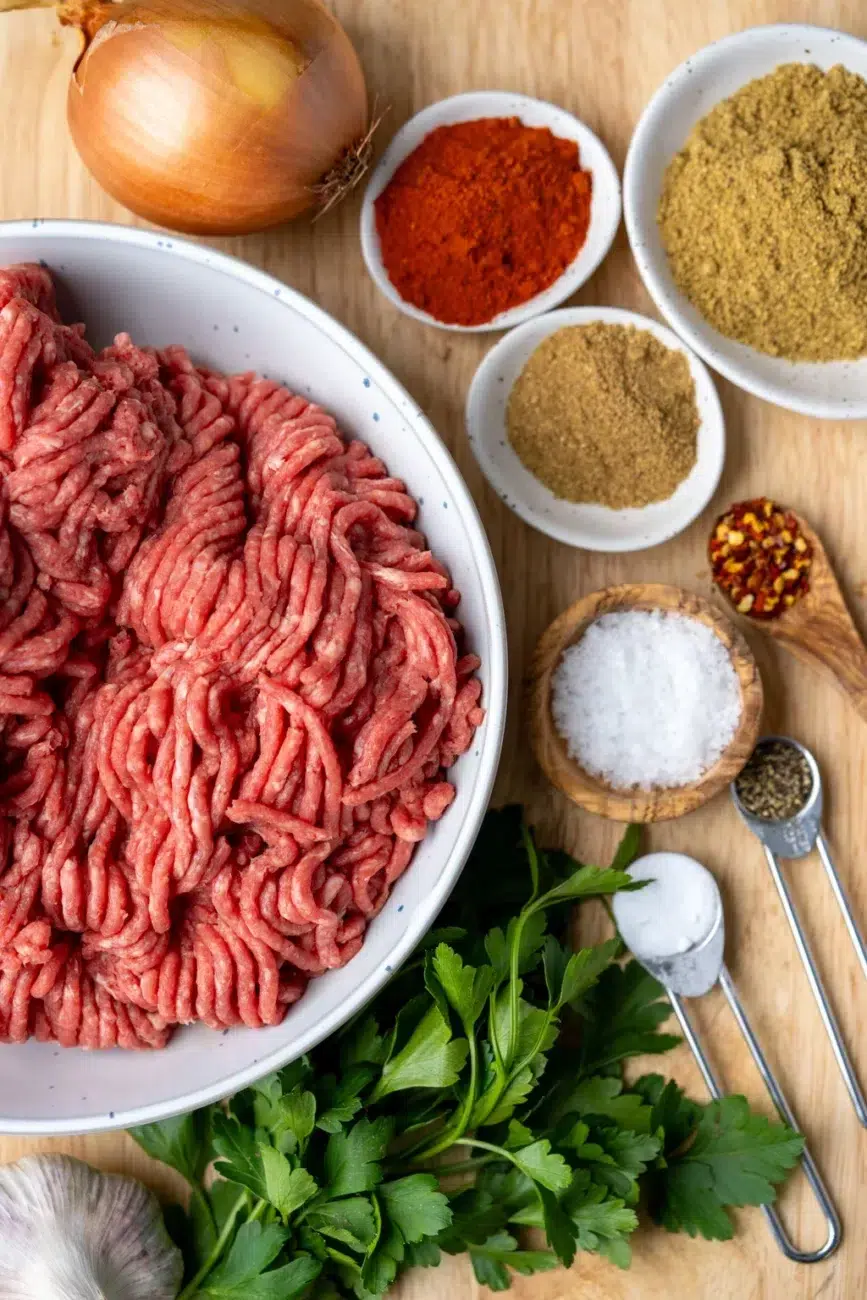 Raw minced meat in a white bowl surrounded by fresh herbs, spices, onion, and garlic – ingredients for juicy oven-baked kebabs