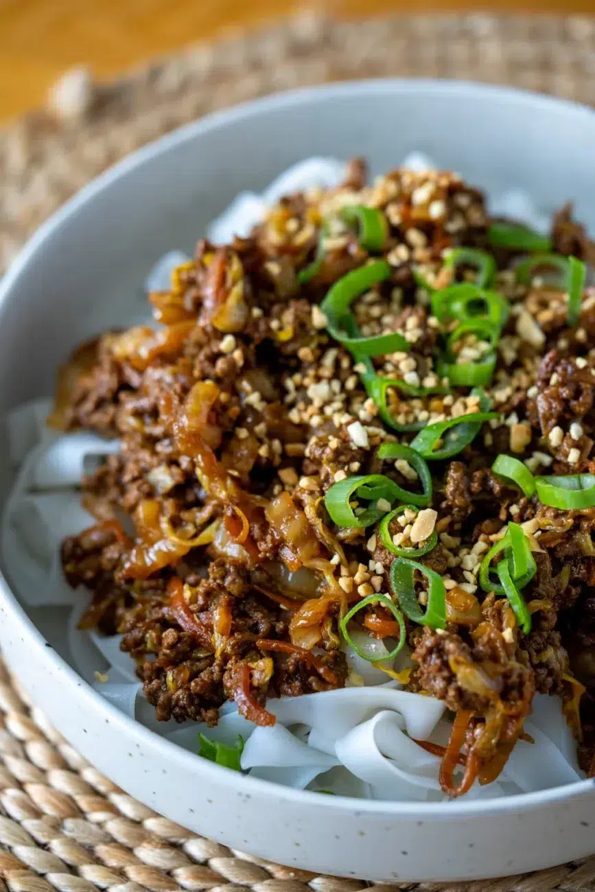 Close-up side view of sticky hoisin beef served over flat rice noodles in a speckled ceramic bowl, topped with chopped green onions and crushed peanuts, set on a woven placemat.
