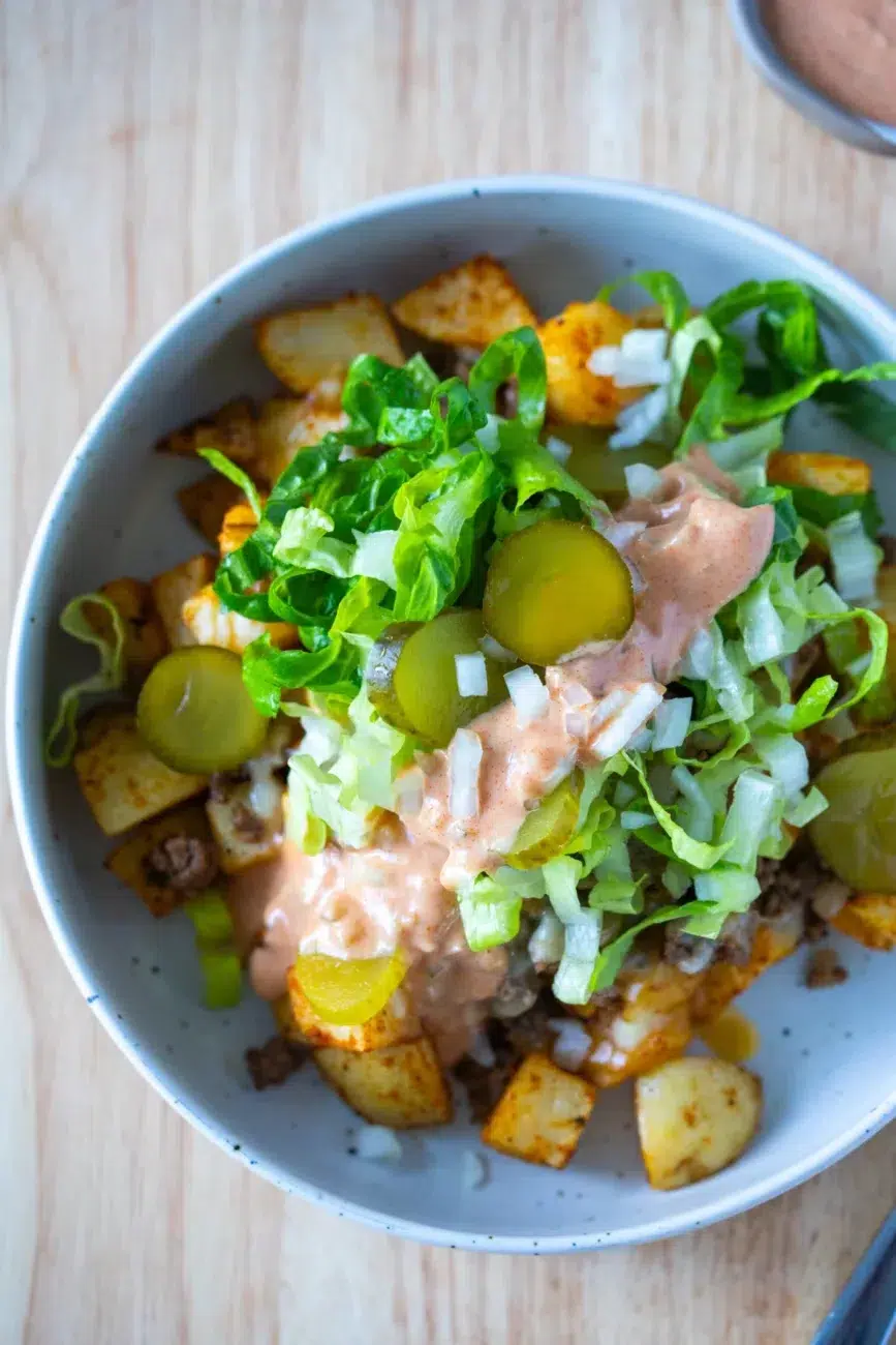 Overhead shot of a Loaded Potato Big Mac Bowl with crispy seasoned potatoes, ground beef, shredded lettuce, sliced pickles, diced onions, and creamy sauce in a white speckled bowl.