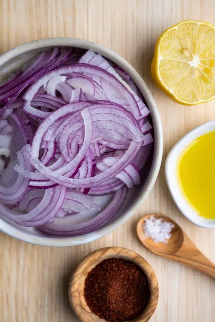 Flat lay of ingredients for sumac onions: sliced red onion, lemon, sumac, olive oil, and salt"