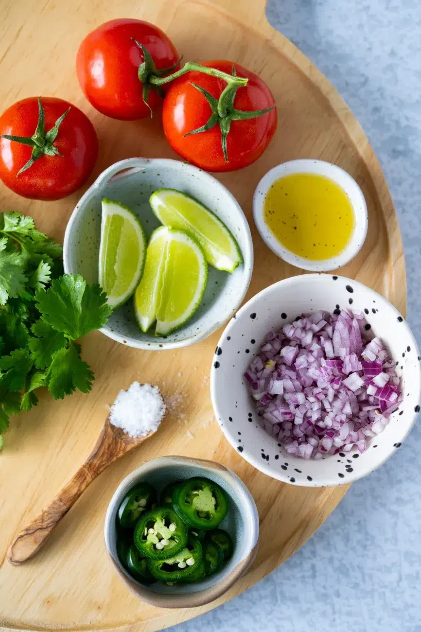 Fresh pico de gallo ingredients on a wooden board, including tomatoes, lime wedges, chopped red onion, sliced chili, coriander, olive oil, and salt