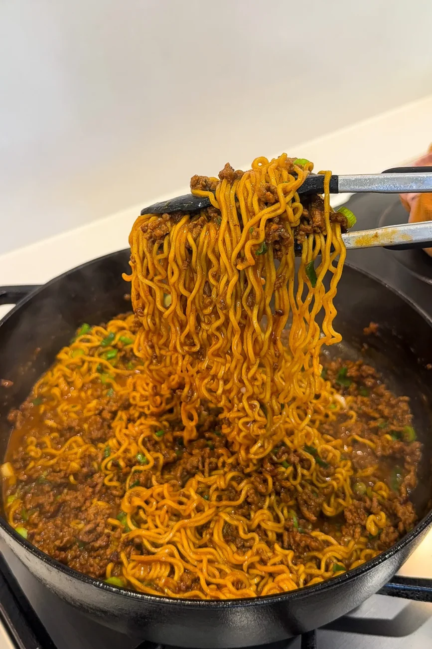 Glossy Korean-style beef noodles being lifted from the pan with tongs
