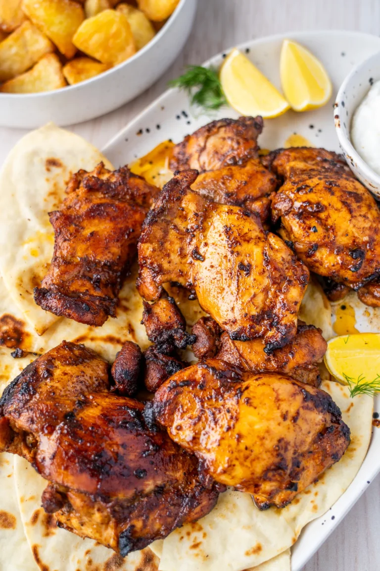 Overhead close-up of golden charred garlic lemon chicken thighs piled on flatbread on a white speckled platter with lemon wedges and crispy roasted potatoes