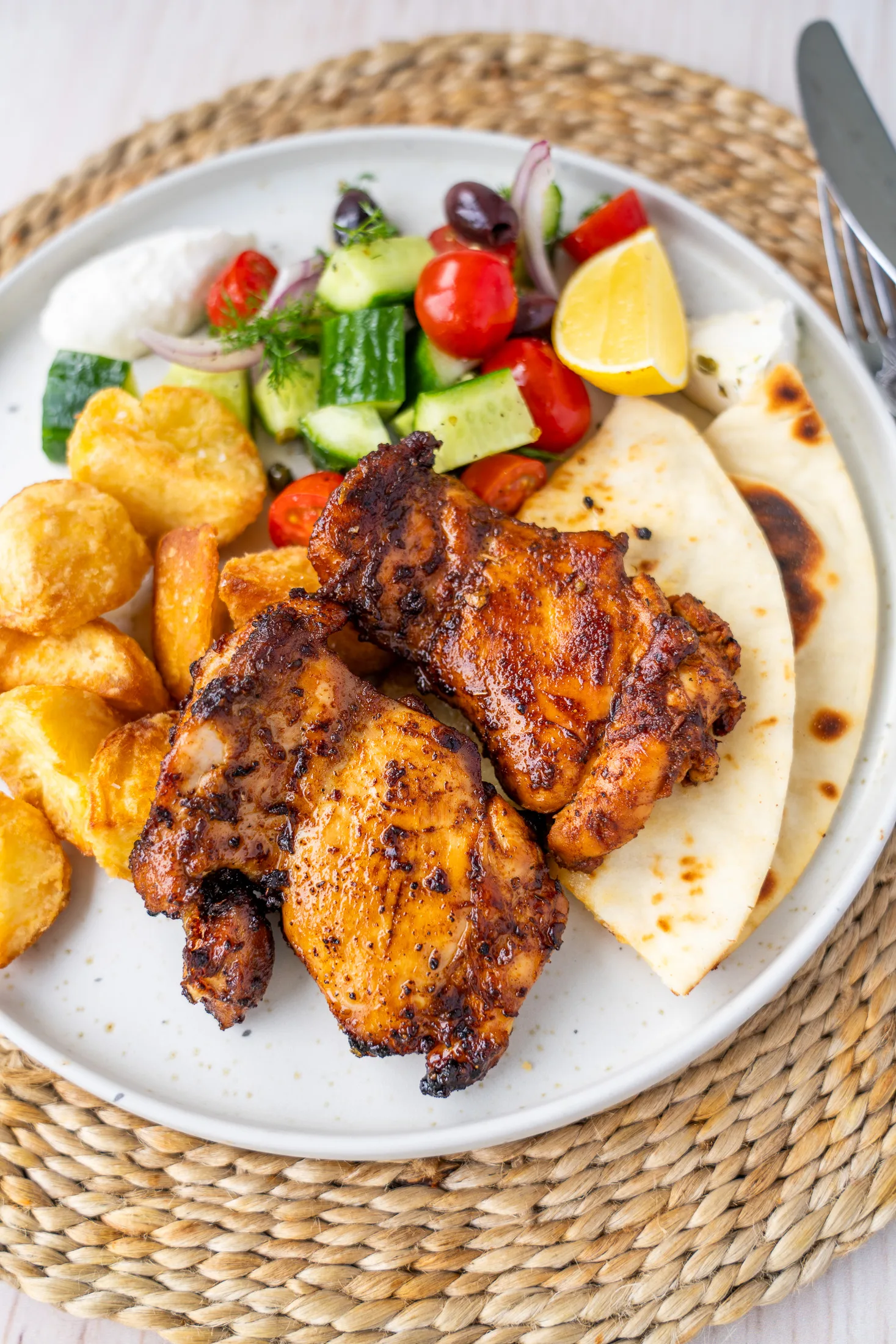 Full dinner plate overhead on a woven placemat with garlic lemon chicken thighs, crispy roasted potatoes, flatbread, village salad with yoghurt and a lemon wedge