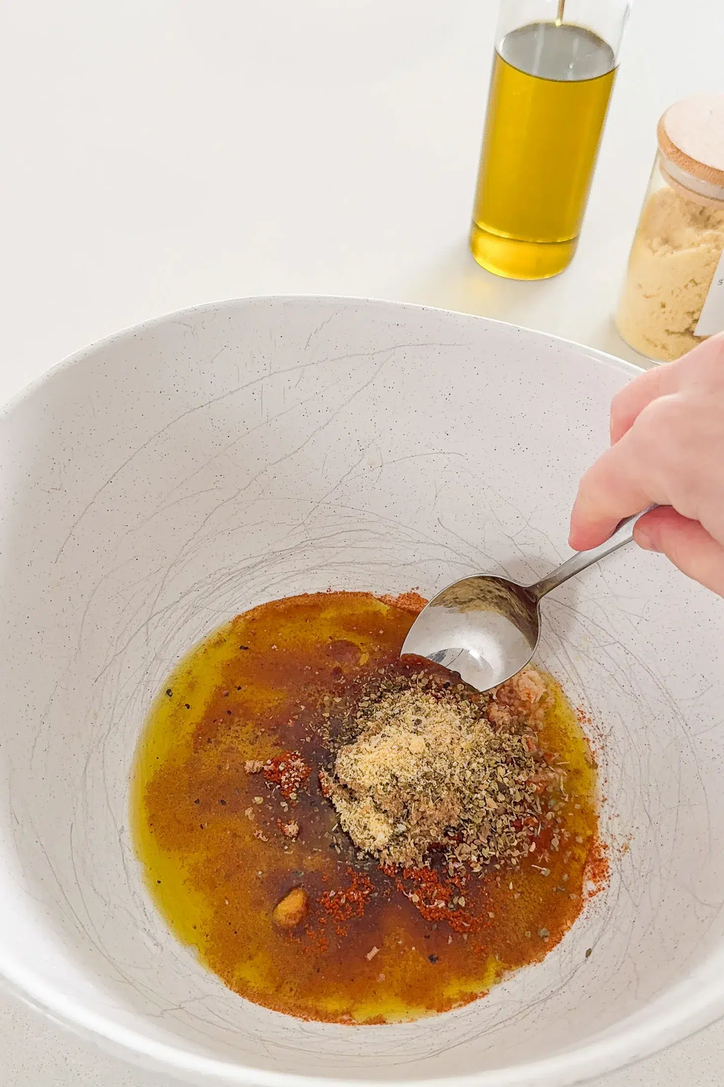 Mixing the garlic lemon marinade in a large white bowl with a spoon, olive oil bottle and garlic powder in the background