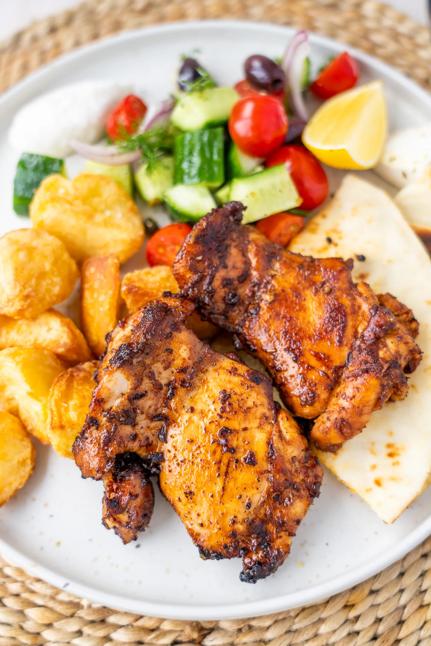 Overhead dinner plate on woven placemat showing garlic lemon chicken thighs, crispy potatoes, flatbread and village salad